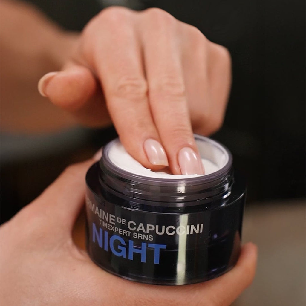 Person holding a jar of skincare product labeled 'NIGHT' with a blurred background